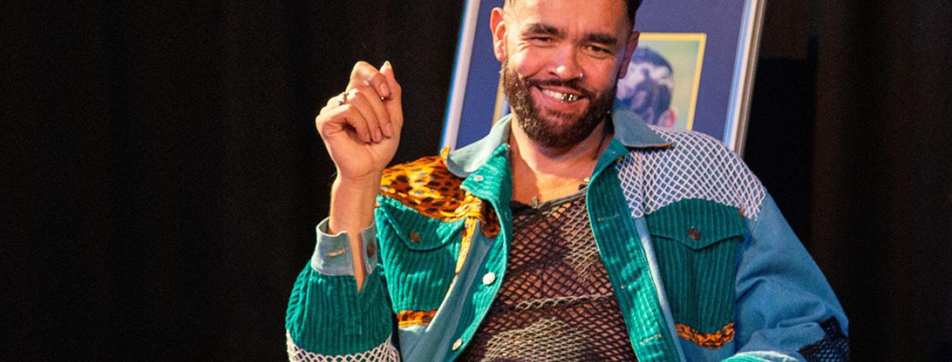  A man with a beard smiles while seated. He wears a colorful outfit and sits next to a stack of fabric. There's a framed picture and a fish decoration in the background.