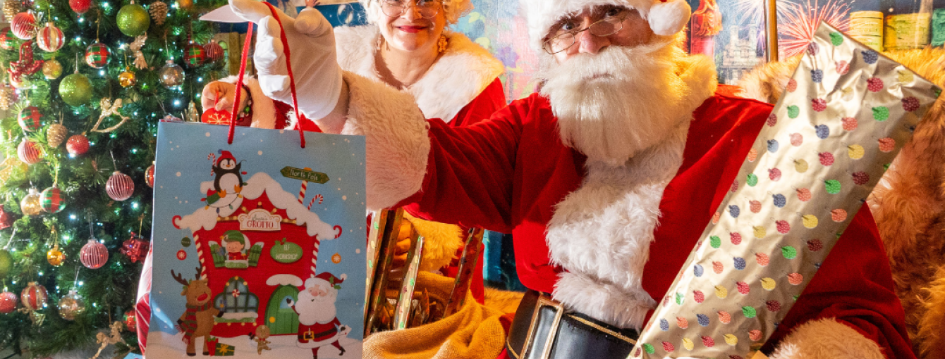 Santa and Mrs Claus sat in a wood cabin next to a decorated Christmas tree. Santa holding two presents and smiling.