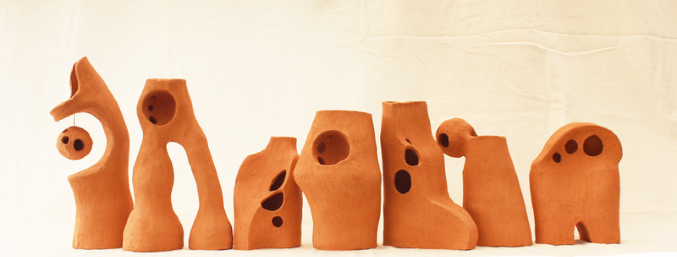 A row of abstract terracotta sculptures with organic shapes, rounded edges, and holes, displayed against a neutral fabric backdrop.