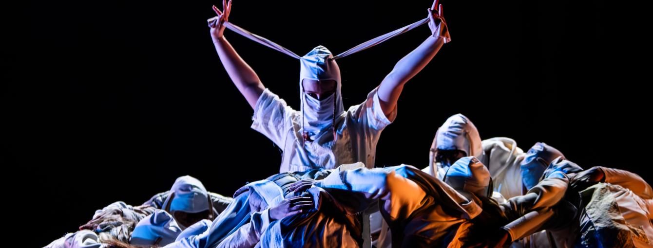 Dancer surrounded by a huddle wears a mask and looks directly at the camera