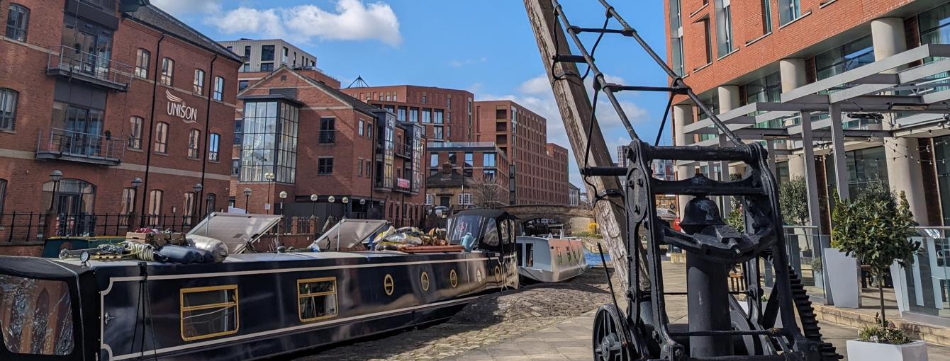 A docked narrowboat next to a heritage crane and surrounded by buildings and blue sky at Granary Wharf in Leeds