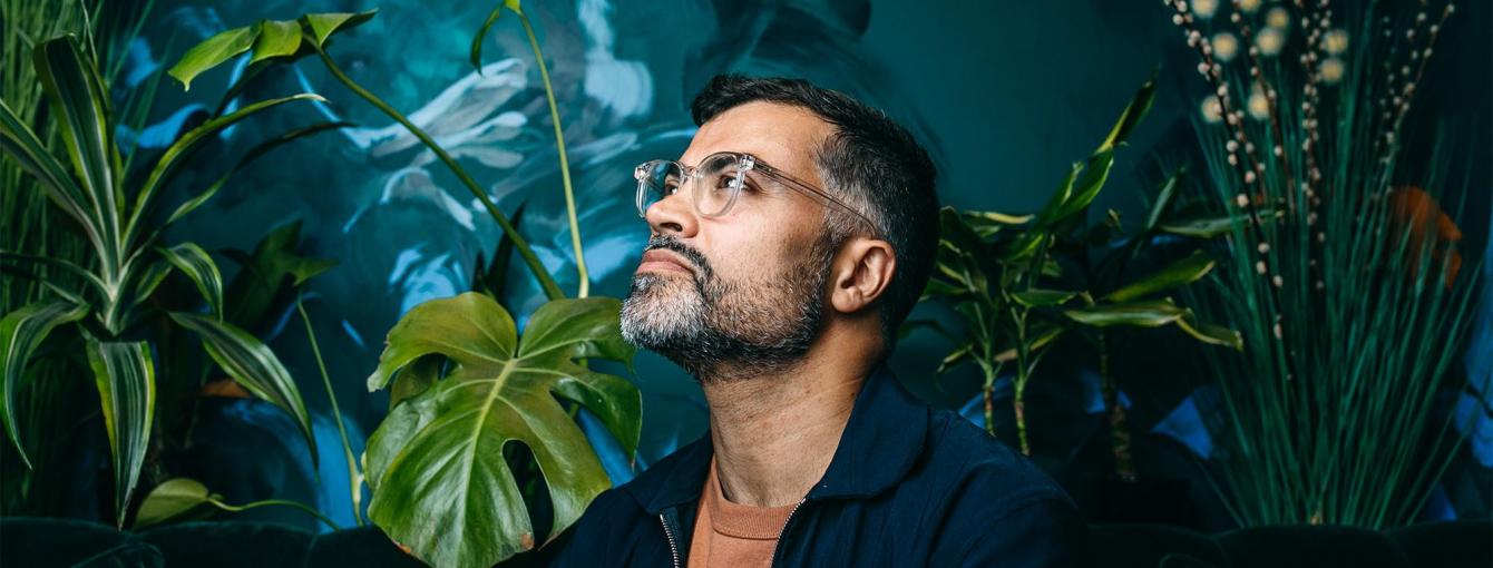 A man wearing glasses and a dark shirt stands in thought in front of a background of plants and vegetation.