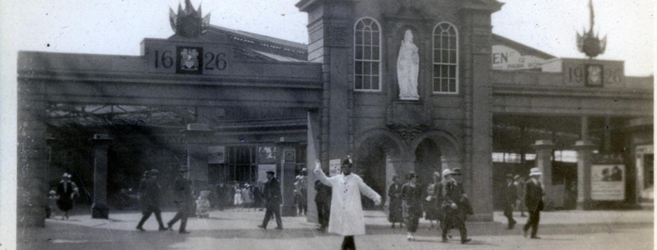 An image of Leeds City Square in 1926, with model moot hall erected for the tercentenary celebrations of Leeds getting its charter. A large building facade with a white statue in a central alcove (Queen Anne) stands in the centre. On either side the dates 1626 and 1926 are etched in what looks like stone. This is entrance to a station concourse. Pedestrians are milling about in period 1920s dress. A policeman in a white coat and gloves stands beneath the arch directing people.