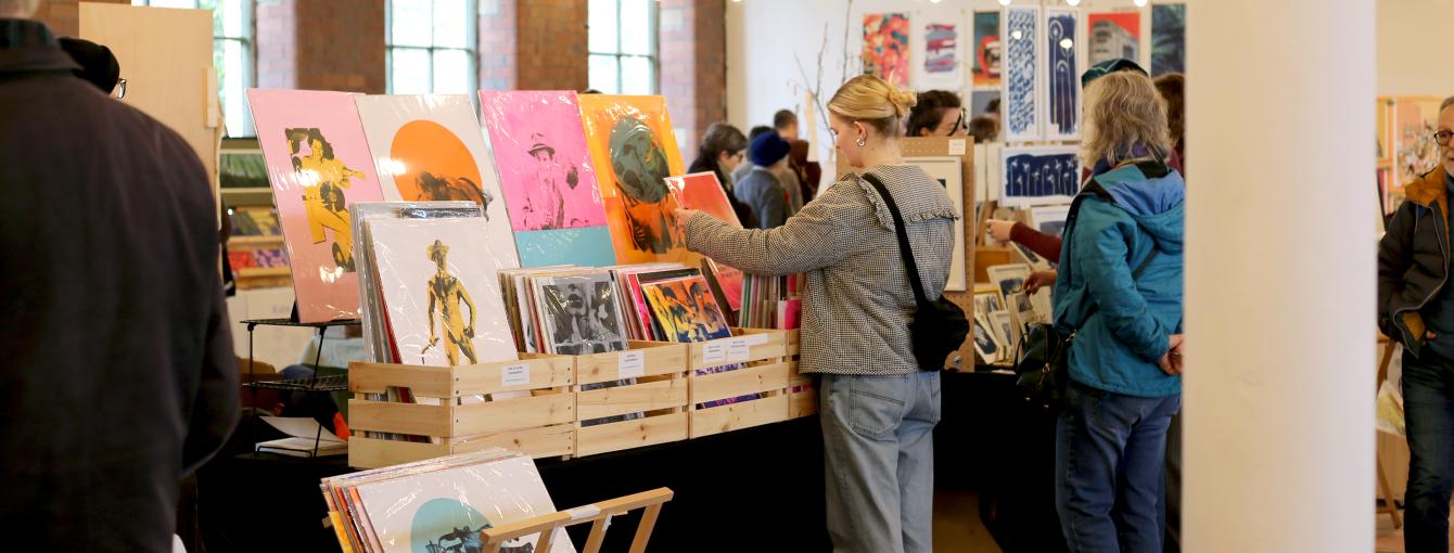 A visitor inside the busy Print Fair at last year's festival inside the 1912 Mill. The space is a former textile mill with red brick walls, white pillars and strings of festoon lights hanging from the high beams. The space is filled with stalls of original prints and handmade books.