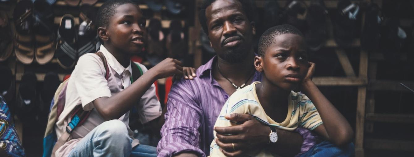 A man sits with two children resting against him while another child sits nearby. They are seated outdoors in front of a display of shoes arranged on shelves.