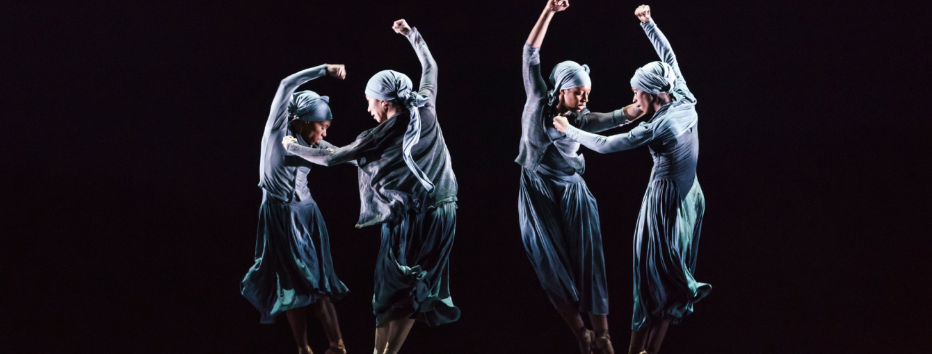 Four performers dance on stage in pairs. They have their arms in the air. 